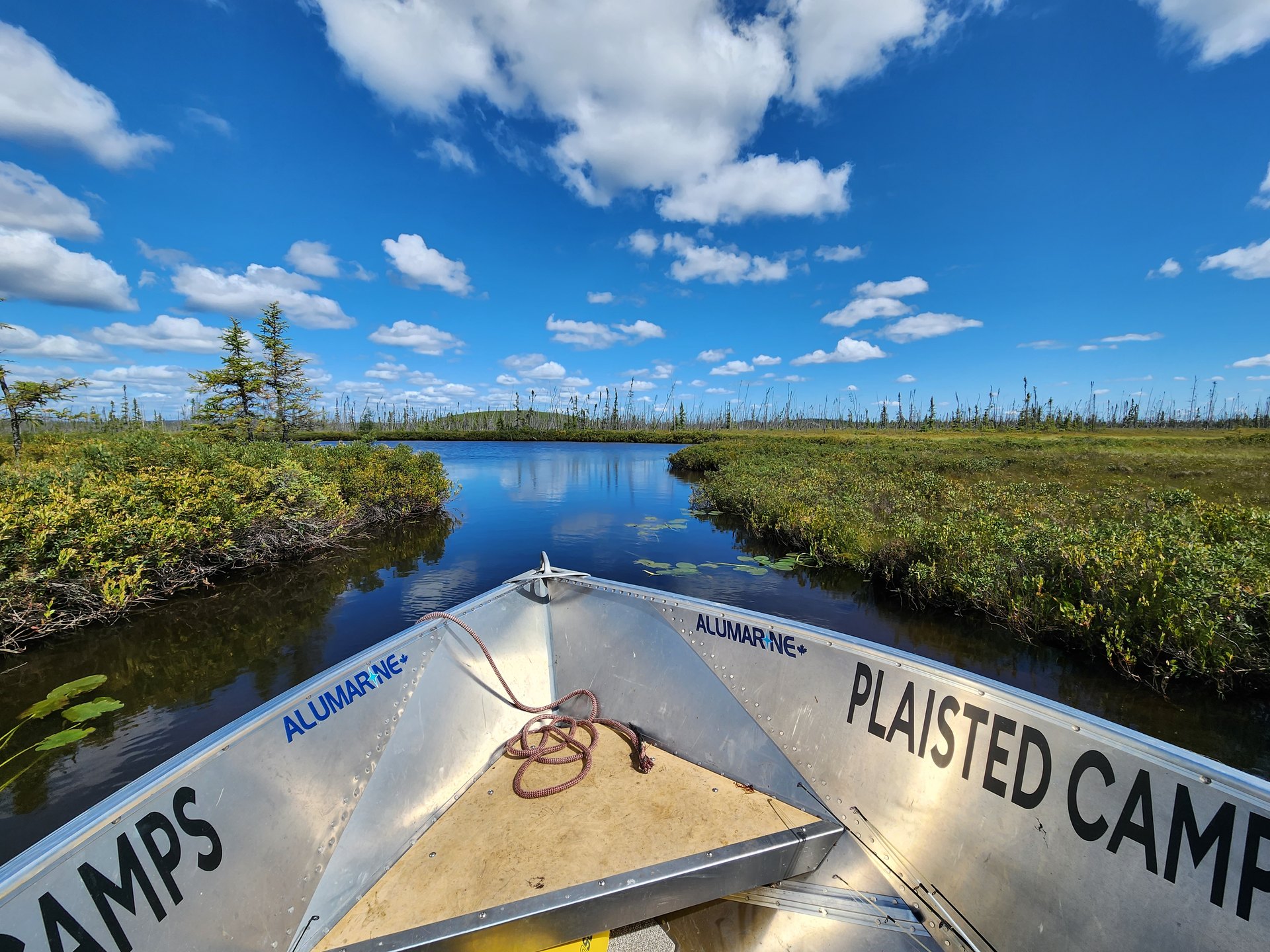 The bow of a Plaisted Camps boat running a narrow channel under a blue sky in northern Saskatchewan